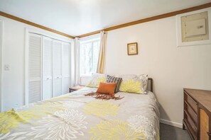 A bedroom with a bed, a window with curtains, a wooden headboard, a bedside table, and a wall-mounted light switch. - Cozy 1-bedroom cottage at Sunnyside with a queen bed, sofa sleeper, and beach access. (Greenbush)