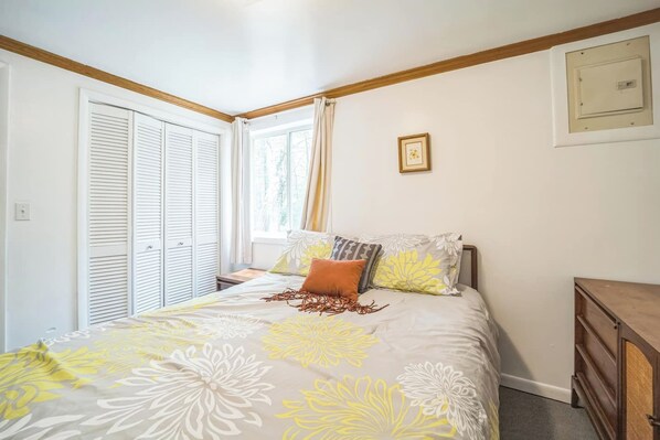 A bedroom with a bed, a window with curtains, a wooden headboard, a bedside table, and a wall-mounted light switch.