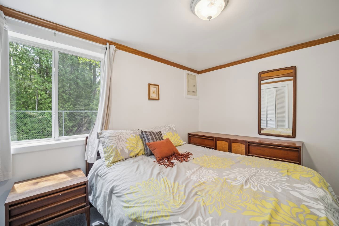 Ein Schlafzimmer mit einem Bett, einem Nachttisch, einem Fenster mit Vorhängen, einem Spiegel und einem hölzernen Kopfteil.