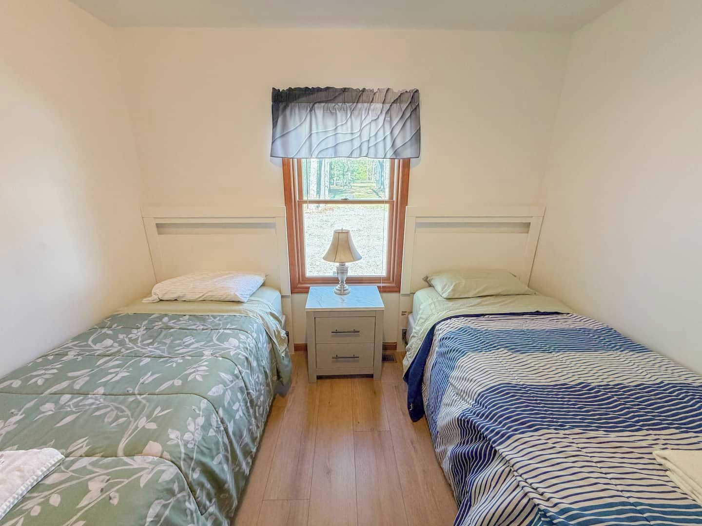 A small, well-lit room with two single beds, a nightstand with a lamp, and a window with a view of greenery outside.