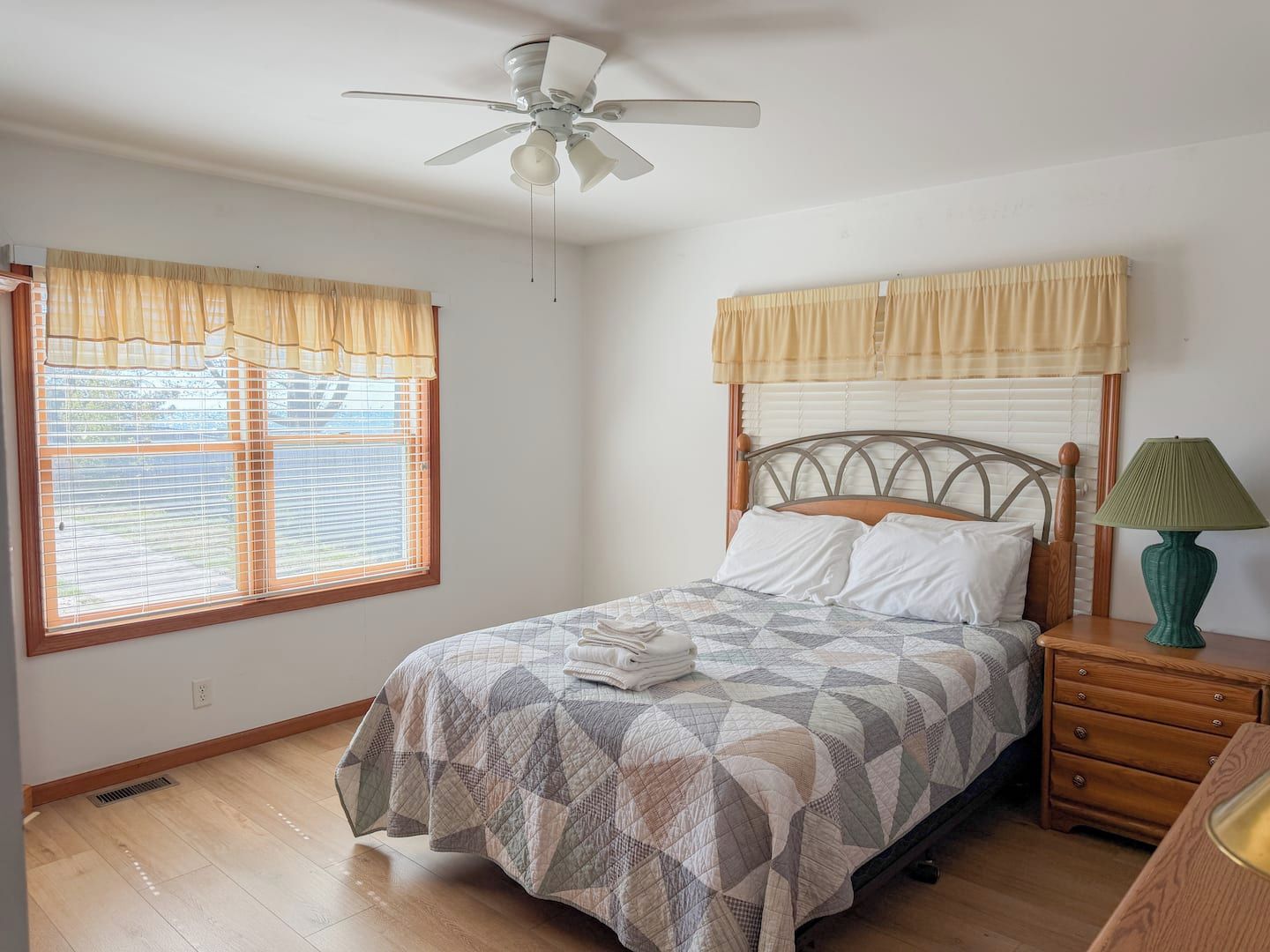 Une chambre à coucher avec un lit, une table de chevet, une lampe, un ventilateur de plafond et des fenêtres avec des rideaux.