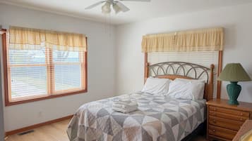 Une chambre à coucher avec un lit, une table de chevet, une lampe, un ventilateur de plafond et des fenêtres avec des rideaux.