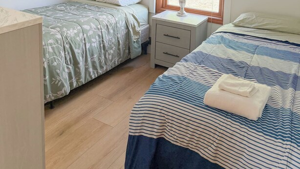 A bedroom with two single beds, a nightstand with a lamp, a window with curtains, and a ceiling light fixture. - The biggest cottage at Sunnyside — newly remodeled, 4BR/3BA, sleeps up to 10. (Greenbush)