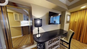 A hotel room with a wooden floor, a black desk with a lamp, a TV mounted on the wall, and a chair.