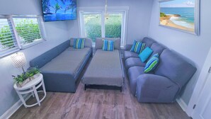 Ein Wohnzimmer mit Deckenventilator, einem Flachbildfernseher, zwei Sofas, einem Couchtisch und einem Fenster mit Jalousien.