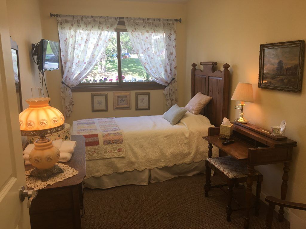 A bedroom with a bed, a desk, a lamp, a chair, a window with curtains, a picture on the wall, and a television on the wall.