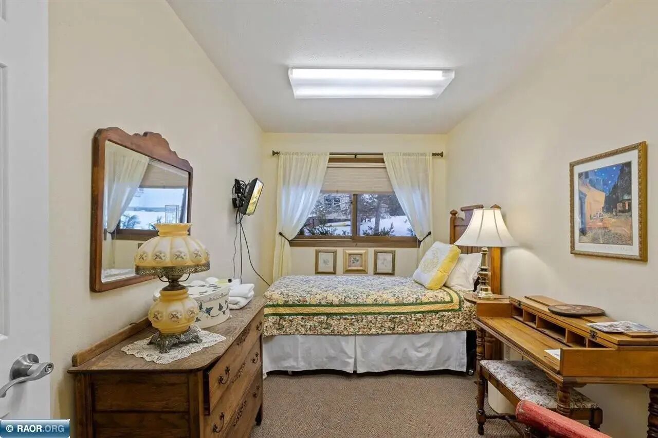 A bedroom with a bed, bedside table, lamp, dresser, and a window with curtains.