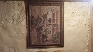 A framed painting of a garden scene with a balcony, mounted on a textured wall. Below the painting, there is a wooden table with two cushions. - Vermilion Park Inn, LL Bear's Den ( lower level) (Soudan)