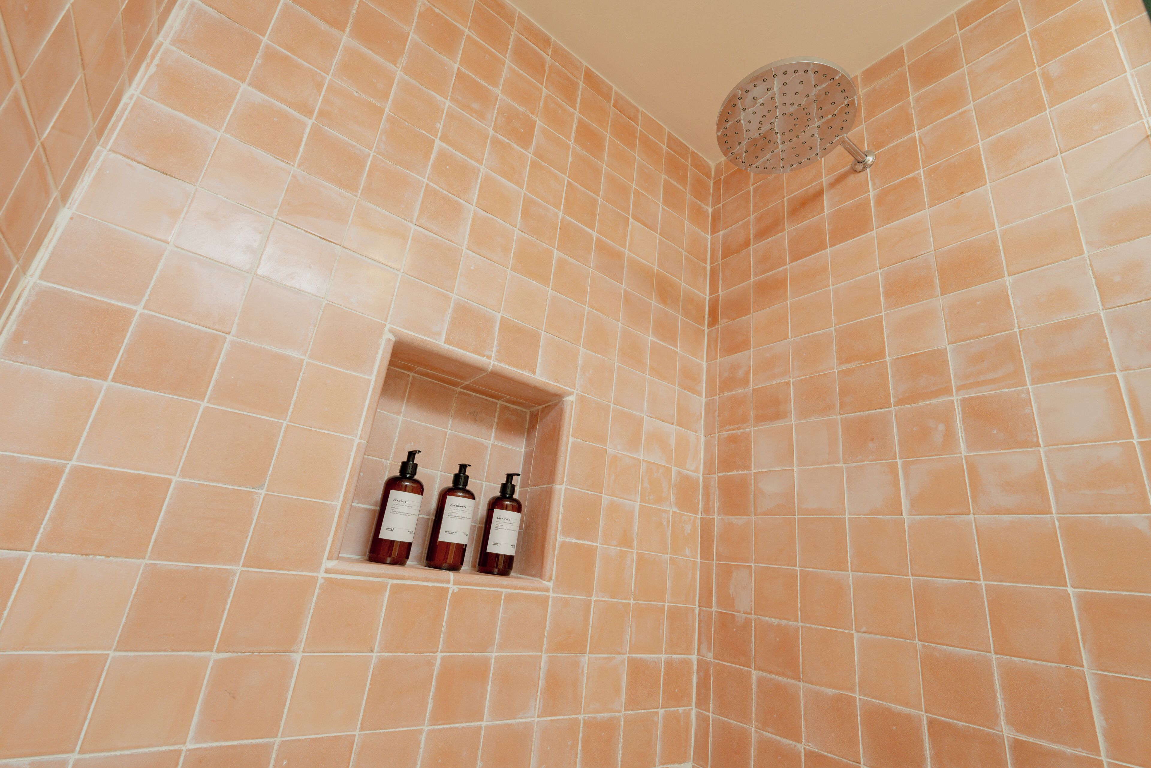 Un baño con baldosas anaranjadas, un cabezal de ducha y tres botellas de champú en un hueco.