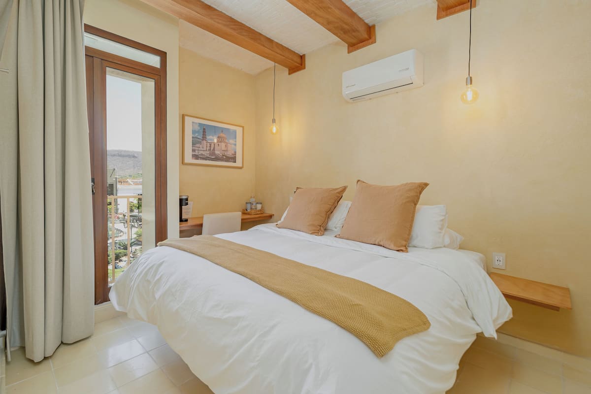 A bedroom with a large bed, beige curtains, a wooden headboard, a nightstand, a painting, and a view through a glass door.