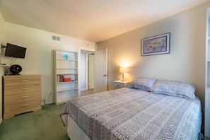 Un dormitorio con cama, cómoda, televisor, estantería y un cuadro en la pared.