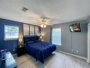 A bedroom with a bed, a ceiling fan, a window with blinds, and a wall-mounted TV.