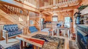 Interior de una cabaña de madera con escalera, sofá, mesa de comedor, zona de cocina y chimenea.