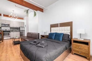 Un dormitorio moderno con una cama grande, cabecera de madera, mesita de noche y vistas a una cocina con microondas y refrigerador.