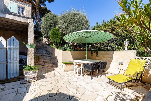 A patio with stone flooring, a table, two chairs, a yellow chair, a green umbrella, and a white fence. - 'Le Repaire' Apartment with Mountain View, Shared Terrace and Garden (Cassis)