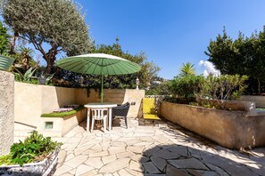 An outdoor patio with stone flooring, a table with chairs, a green umbrella, and a yellow chair. - 'Le Repaire' Apartment with Mountain View, Shared Terrace and Garden (Cassis)