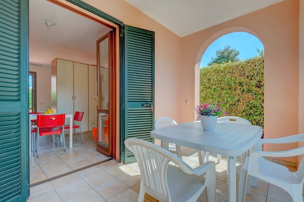 An open door leading to a dining area with a table and chairs, a potted plant, and a view of a hedge outside.