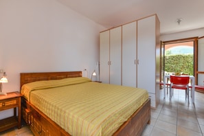A bedroom with a bed, a wardrobe, a desk with a lamp, and a view of greenery outside.