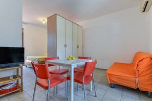 A room with a dining table, four red chairs, a white cabinet, a television, a covered orange sofa, and a bed with a yellow bedspread.