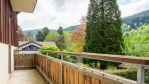 A balcony with a wooden railing, a view of a forested area, and a building with a sloped roof in the background. - Apartment 'Des Sapins' with Mountain View, Private Terrace and Wi-Fi (La Bresse, Grand Est, France)