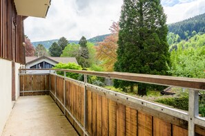 A balcony with a wooden railing, a view of a forested area, and a building with a sloped roof in the background.