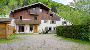 A two-story wooden building with a balcony, a gravel driveway, and a forested hillside in the background. - Apartment 'Des Sapins' with Mountain View, Private Terrace and Wi-Fi (La Bresse, Grand Est, France)