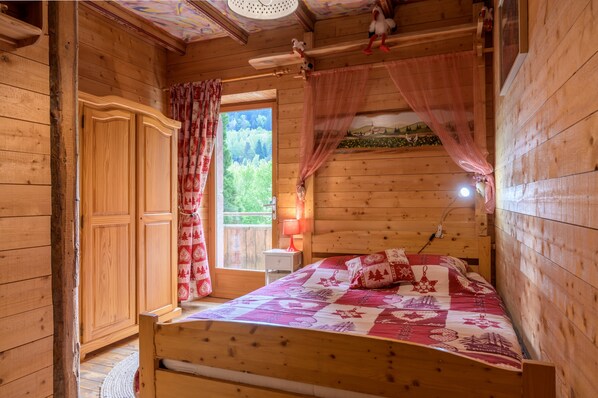 A wooden cabin bedroom with a bed, a wardrobe, a window with curtains, and a small table with a lamp.