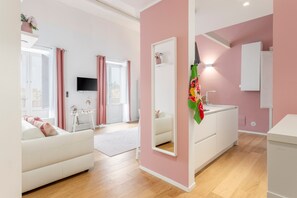 Un salon moderne avec un canapé blanc, un mur rose, un grand miroir et un coin cuisine doté de meubles blancs.