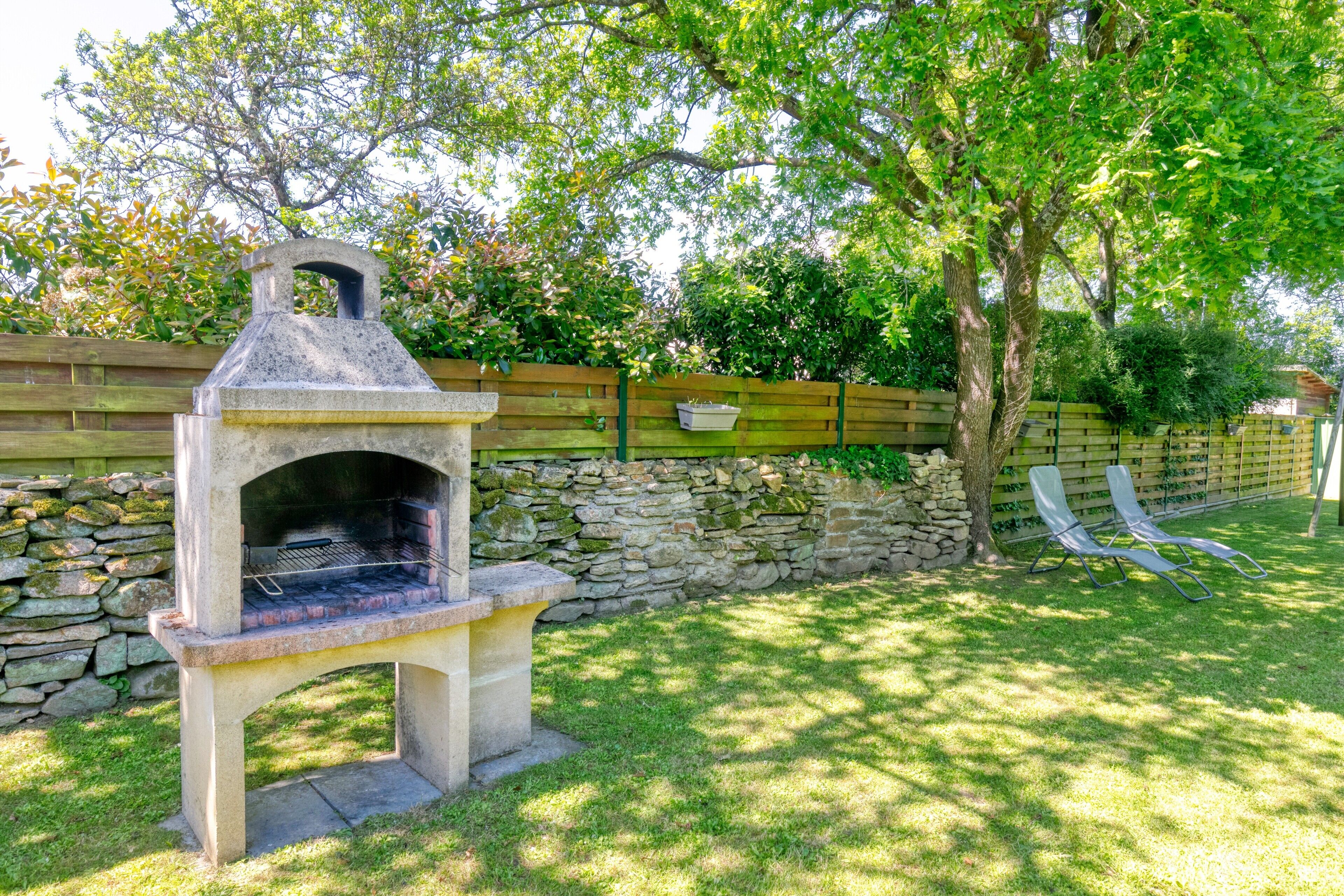 Una griglia in pietra in un giardino sul retro, con un muro in pietra, una recinzione in legno e due poltrone da relax.