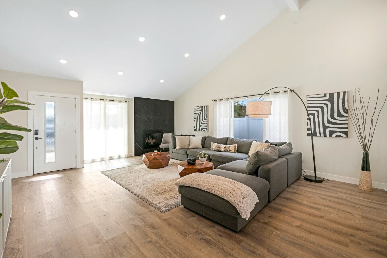 Ein modernes Wohnzimmer mit grauem Sofa, einem Couchtisch und einer Stehlampe. Links ist eine Tür und eine schwarze Wand mit Kamin vorhanden.