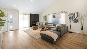 Ein modernes Wohnzimmer mit grauem Sofa, einem Couchtisch und einer Stehlampe. Links ist eine Tür und eine schwarze Wand mit Kamin vorhanden.