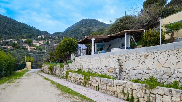 A rural road with stone walls, a covered outdoor seating area, and a mountainous backdrop.