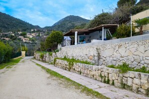 Une route de campagne bordée de murs en pierre, un espace extérieur abrité avec des sièges, et un paysage montagneux en arrière-plan.