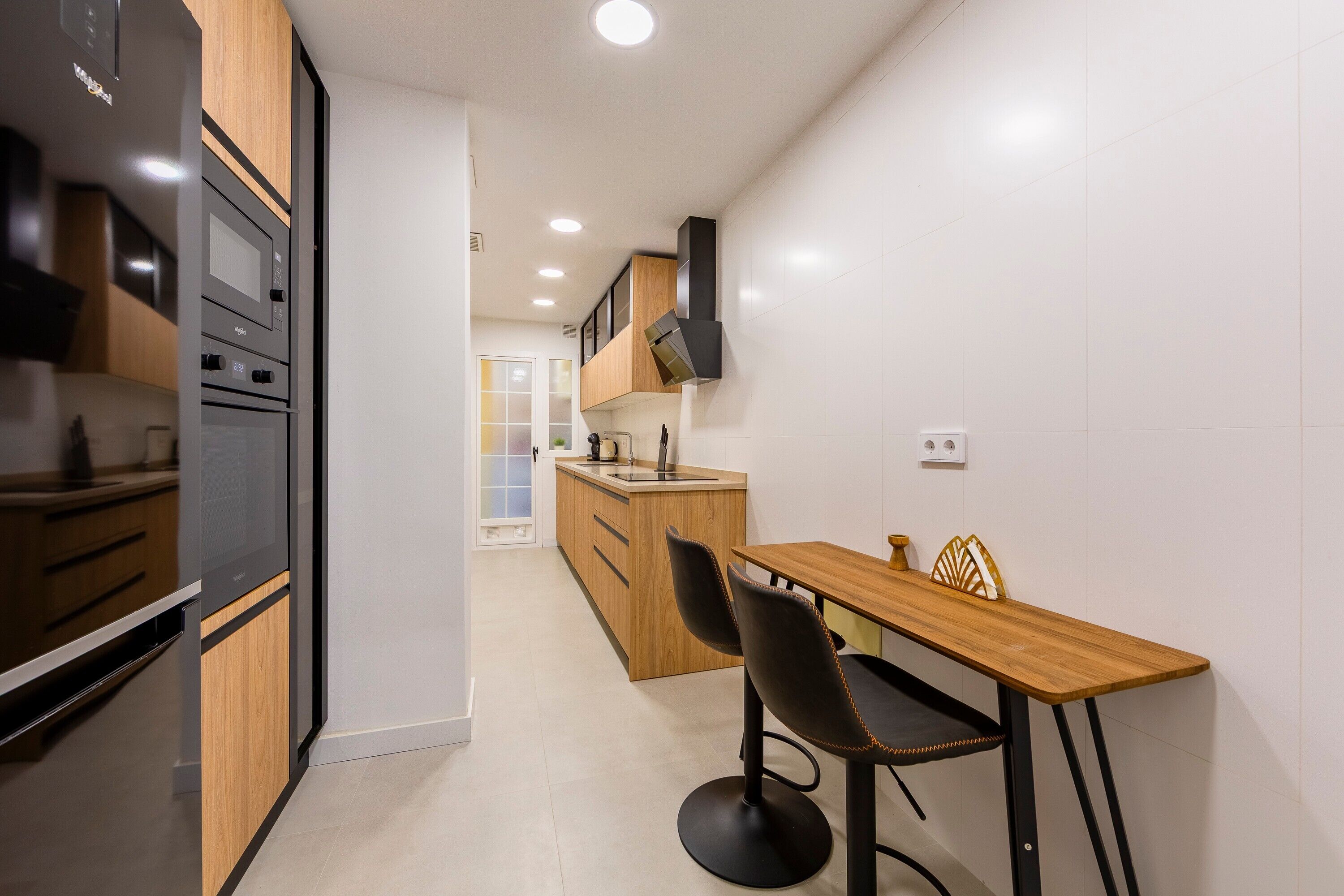 A modern kitchen with built-in appliances, a wooden dining table with two chairs, and a view into the adjacent living area.