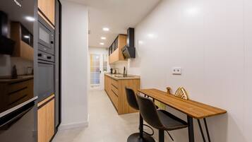A modern kitchen with built-in appliances, a wooden dining table with two chairs, and a view into the adjacent living area.