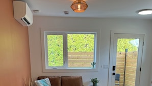 Una sala de estar con un sofá marrón, una mesita auxiliar, una ventana con vistas a la vegetación y una puerta que da al exterior.