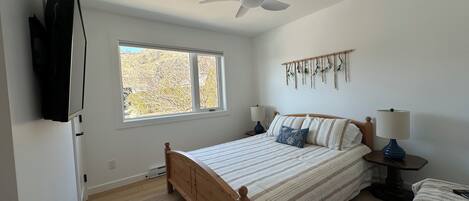 A bedroom with a bed, a ceiling fan, a window, a wall-mounted TV, and a bedside table with a lamp.