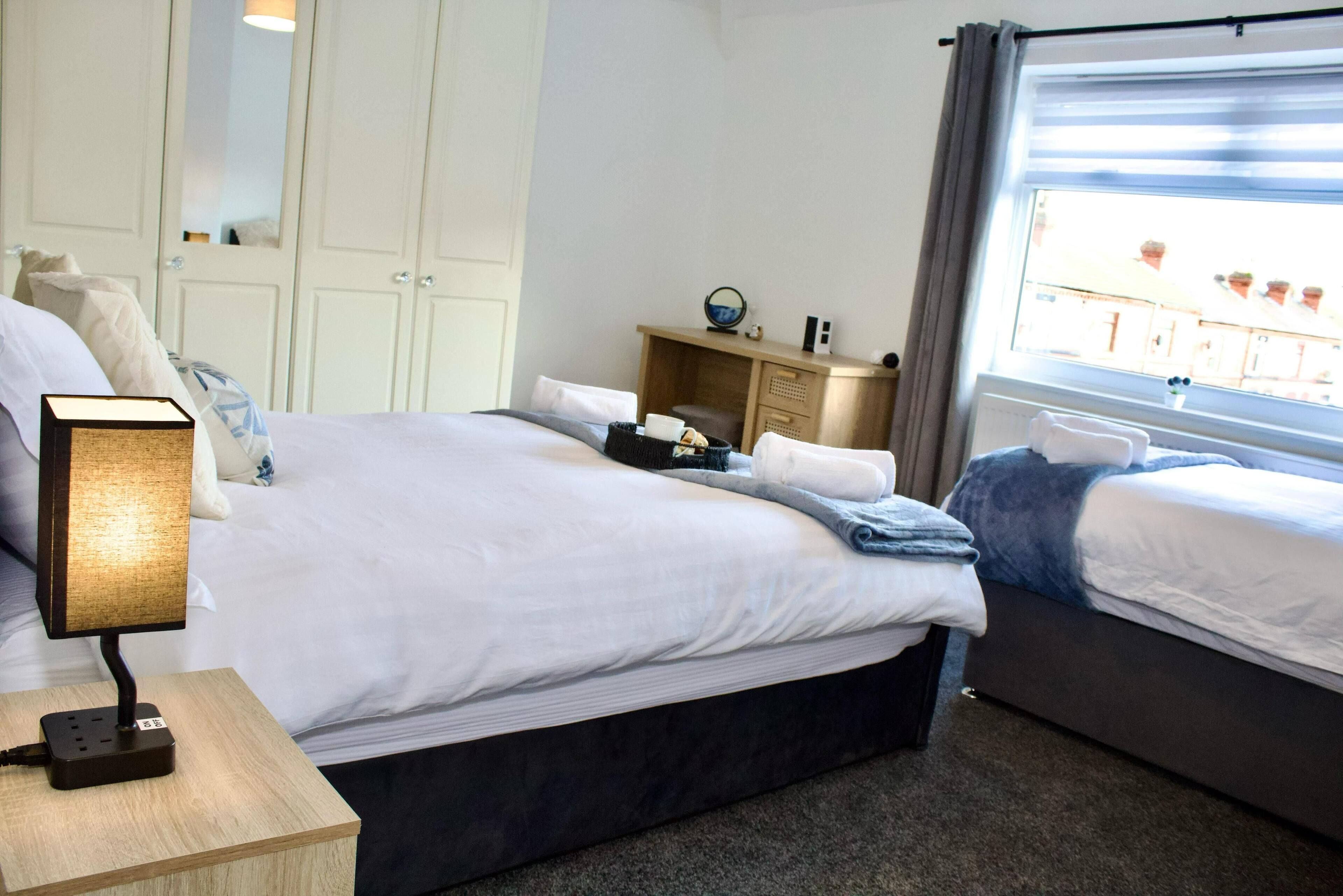 A hotel room with a large bed, a bedside table with a lamp, a small desk with a mirror, and a window with a view of buildings.
