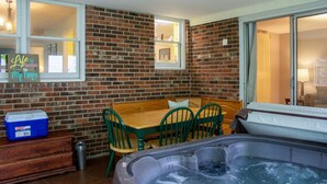 A cozy outdoor seating area with a brick wall, a wooden table, and green chairs. A hot tub is visible in the foreground. - Huge Manor w Indoor Hot Tub and Pool Table (Bloomington)