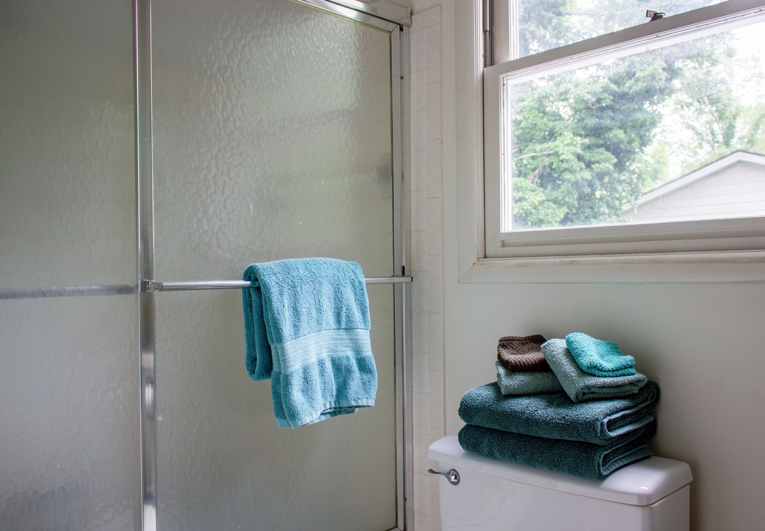 A bathroom with a glass shower door, a towel rack with a single towel, and a stack of folded towels on the toilet.