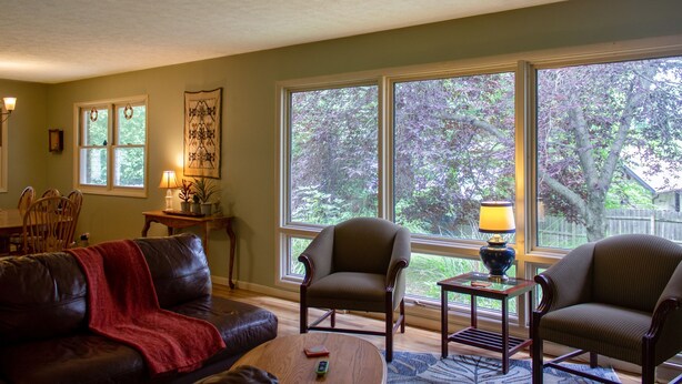 A living room with a leather sofa, two armchairs, a coffee table, and a lamp. There are large windows with a view of trees. - Huge Manor w Indoor Hot Tub and Pool Table (Bloomington)