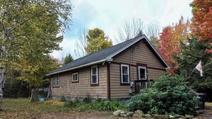 A single-story house with a gabled roof, a front porch, and a small front yard with a bush and a stone wall.
