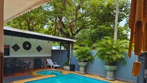 Uma piscina com deck revestido, rodeada por plantas em vasos e uma área de estar coberta.