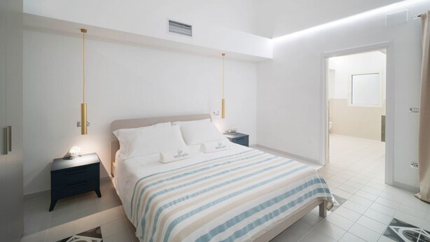 A modern bedroom with a bed, bedside tables, and a door leading to another room. - WePuglia - The White Luxury – Apt Ataraxia (Monopoli)