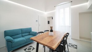 A modern living room with a wooden dining table, a blue sofa, and a large window with sheer curtains. - WePuglia - The White Luxury – Apt Ataraxia (Monopoli)