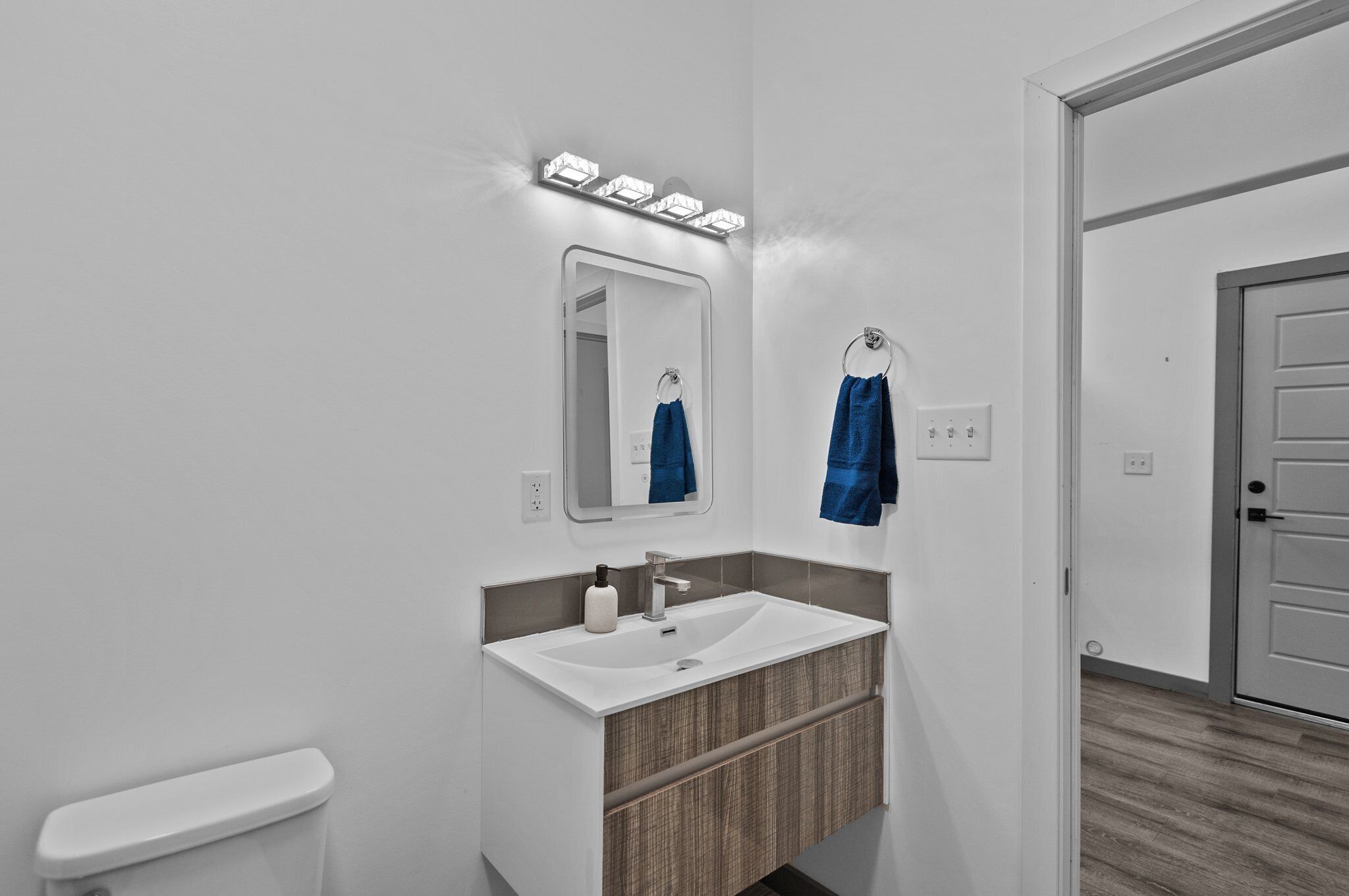 A modern bathroom with a white sink, mirror, and toilet. There are blue towels hanging on hooks, and a lighted vanity area.