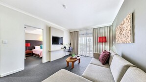 A hotel room with a living area, a bed, a TV, a small table, and a window with curtains. - Resort Style City View w/ King Bed, Pool, Balcony (Darwin City)