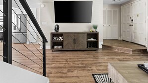 Een moderne woonkamer met een grote flatscreen televisie aan de muur, een visgraat parketvloer, een donker houten tv-meubel en een trap met zwarte leuningen.
