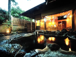 A traditional Japanese onsen with a wooden exterior, a stone hot spring, and a wooden fence in the background.