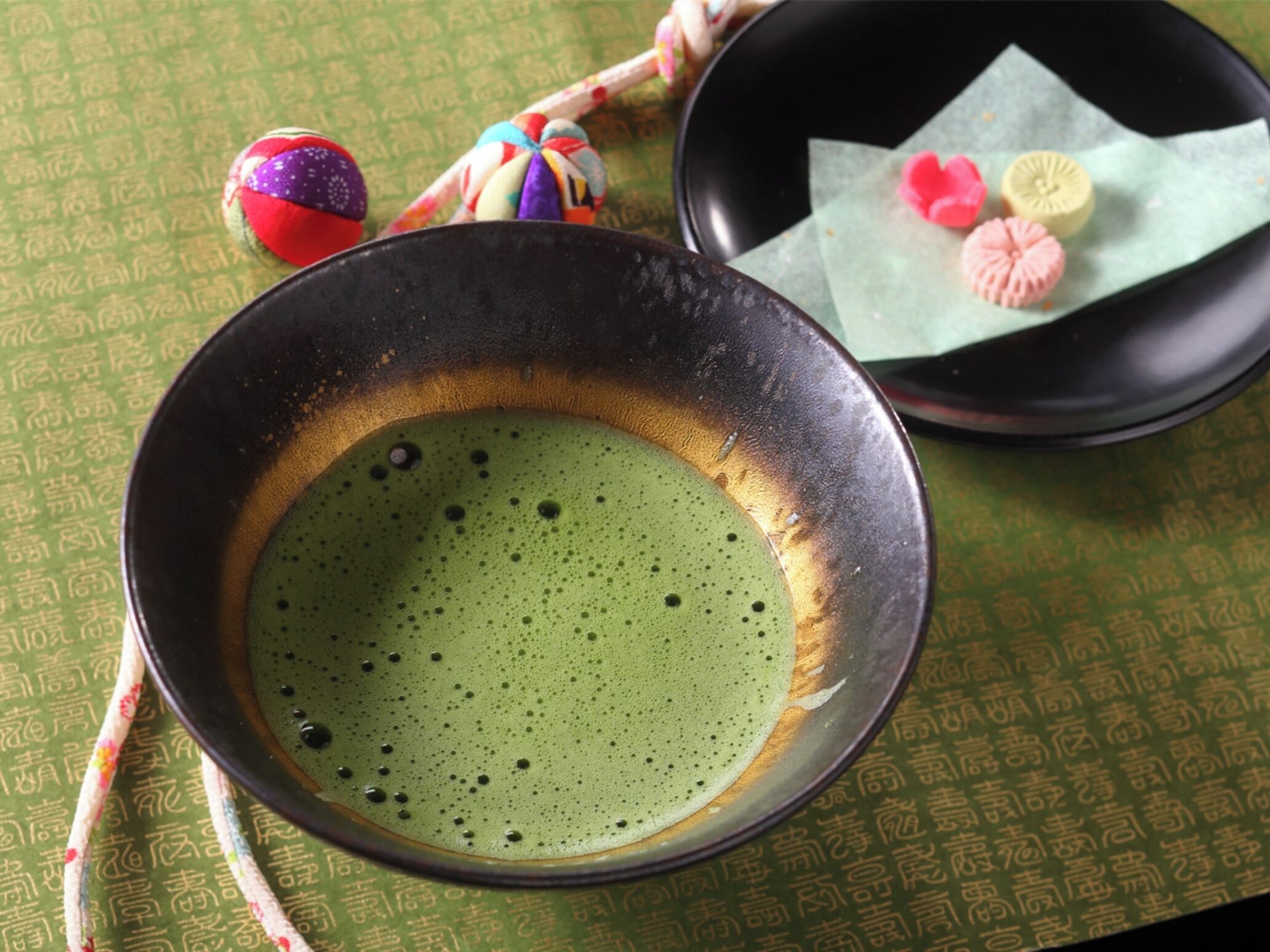 A bowl of green tea with bubbles, a black bowl, and a plate with pink and yellow items on a green patterned tablecloth.